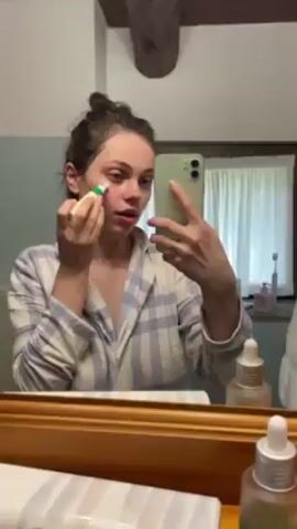 A young woman in blue and white striped pajamas is looking at herself in a mirror while applying a skincare product to her brow bone using a rolling applicator. She is holding her phone in her other hand to record the application. The bathroom mirror is framed with wood and sits above a counter with several skincare bottles and a toothbrush holder. A window with sheer white curtains is visible in the background. The woman’s hair is pulled up into a loose bun.
