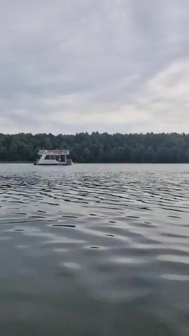 A white boat with a blue stripe moves across a calm lake. The boat has two levels and several people are visible on the top deck. A line of dark green trees forms the background. The sky is cloudy and overcast. The water has gentle ripples.