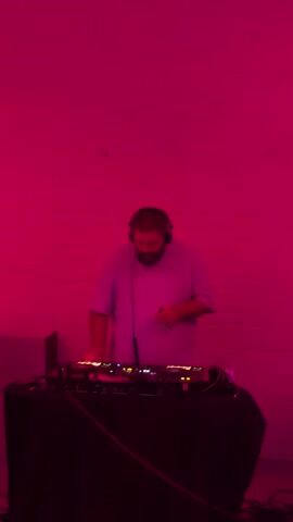 The video shows a DJ performing at a venue. The DJ is a man with a beard, wearing headphones and a light-colored t-shirt. He is standing behind a DJ console with turntables and a mixer, with various cables connected to the equipment. The setting appears to be an indoor space with a white brick wall in the background. The room is illuminated with a vibrant pink light, creating a dramatic and atmospheric mood. The DJ is actively engaged with the equipment, with his hands on the turntables and mixer, suggesting he is in the process of playing music. The video captures him adjusting his headphones and interacting with the music. The overall impression is of a lively event, likely a party or club night. There is no visible text in the video. The time of day and weather are not discernible from the footage, but the indoor setting and artificial lighting suggest it could be evening or nighttime.