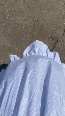 A person wearing a blue and white striped dress and brown sandals walks on a concrete surface. The dress has a ruffled collar. The camera is positioned to film the person's feet and the lower part of the dress as they walk away. The sun casts a shadow of the person on the ground.