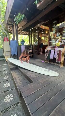 A woman with curly brown hair wearing a black one-piece swimsuit is crouching on a wooden platform, applying something to a long surfboard. She is barefoot and wearing a watch on her left wrist. The surfboard is white and has a stripe down the center. Behind her, on the same platform, are clothing displays and inside the building behind that, a man is seen near the entrance of the shop. The platform is part of the shop's exterior and has decorative cobblestone pathways leading up to it. The shop appears to be located in a tropical or subtropical environment, based on the vegetation. The lighting suggests it is daytime.