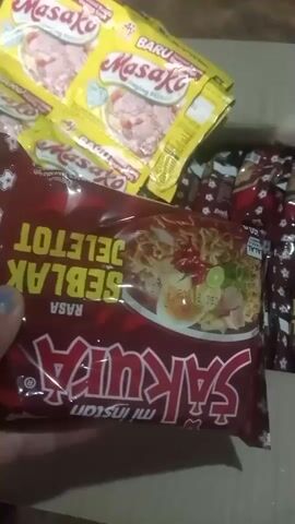 The video displays a close-up shot of several packages of instant noodles. In the foreground, a hand is holding a dark red package of "Sakura" brand instant noodles with "Mi Instan" written below it. The flavor visible on the package is "SEBLAK JELETOT". Behind this package, several yellow packages of "Masako" brand seasoning are visible, some with "BARU" indicating new. To the right, partially obscured, are more packages of what appear to be instant noodles, with a red and white striped pattern. The background is dark, suggesting an indoor setting, possibly a box or table. The lighting is dim. The video is short, with the spoken content being a brief endorsement of the noodles.