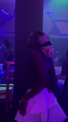 The video shows a woman with blonde braided hair tied up dancing in a club. She is wearing a black top and a white ruffled skirt. The club is dimly lit with purple and green lights. There are other people in the background, also dancing. The woman turns her back to the camera and dances. The video pans slightly to show more of the club environment and other patrons.