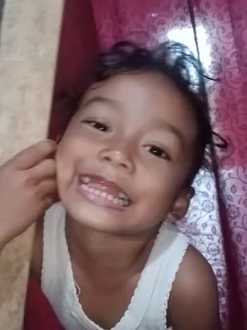 This media file captures a close-up view of a young child lying down. The child, with dark hair and brown eyes, is wearing a white tank top. They are looking directly at the camera with a wide, open-mouthed smile, revealing their teeth. The child's facial expression conveys happiness and playfulness.

The background consists of a textured, pink fabric with a floral pattern, possibly a bedsheet or curtain. A wooden object, which appears to be a plank or stick, is partially visible in the foreground, held by the child's hands. The lighting suggests an indoor setting, possibly during the day, as indicated by a bright light source reflecting on the background.

The child's posture, with arms extended and head tilted back, suggests they are engaged in some form of play or interaction. The close framing of the child's face emphasizes their joyful expression. The overall mood is lighthearted and cheerful. There is no visible text in the media file. The location context is Cotseutui, Indonesia.