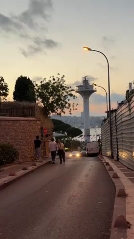 The video shows a street scene at dusk. The street slopes downwards and is lined with stone walls and a metal fence. Several people are walking up the street. A white van and a car with headlights on are driving down the street. In the background, there is a body of water and a tall white tower with a light on top, resembling a lighthouse. The sky is cloudy with hints of pink and orange from the setting sun. Streetlights are on.