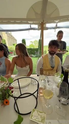 The media shows a wedding reception taking place under a white tent. The setting is outdoors, with views of a building and greenery visible through the tent's clear walls, suggesting a rural or countryside location in the Cotswolds.

Several tables are set for a meal, adorned with floral centerpieces, wine bottles, and place settings. People are seated at the tables, dressed in formal attire, indicating a celebratory event. The subjects include a bride in a white strapless dress and a groom in a yellow vest and tie, seated at the head of a table. Other guests are visible, some engaged in conversation or looking towards the camera. Waitstaff are also present, one of whom is carrying a platter of food.

The lighting suggests it is daytime, possibly late afternoon, with natural light filtering into the tent. The overall atmosphere appears festive and convivial. A small, handwritten place card is visible on the table with the name "Jack m." The video captures a dynamic scene with some panning and movement, creating a sense of lively activity.