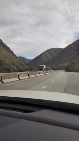 A dashboard view from inside a vehicle shows a paved road with two lanes of traffic, separated by white lines. The road curves to the right as it travels through a mountainous landscape. The terrain on either side of the road is characterized by steep, rocky hillsides covered in sparse vegetation. A concrete barrier separates the road from the slope on the left. Two trucks are visible on the road ahead, one a red and white tanker truck in the left lane, and another truck further ahead in the right lane. The sky is overcast with clouds.