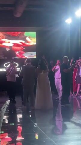 A wedding ceremony is taking place indoors with a wedding couple standing on a shiny black dance floor facing away from the camera. They are both dressed formally, with the bride wearing a white gown and the groom in a grey suit. There is a large screen behind them displaying an image of a wedding cake. To their left, a man in a white t-shirt and black pants is operating a keyboard. To their right, a crowd of people are watching and taking pictures with their phones, with some individuals also holding microphones. The environment appears to be a reception hall with dramatic lighting from the ceiling. The floor reflects the lights and the people, creating a glossy effect.