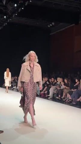 A blonde female model walks down a runway at a fashion show, wearing a beige leather jacket over a long, patterned dress with a slit. She carries a dark purse in her left hand and wears high-heeled shoes. The runway is white, and the background shows an audience seated in tiered seating, illuminated by stage lights. The model turns her back to the camera as she walks away.