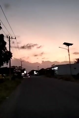 The video shows a straight, paved road lined with buildings and utility poles. The sky is a gradient of pale orange and grey, suggesting dusk or dawn. Light from buildings and streetlights is visible along the road. A single motorcycle is seen driving away in the distance. The scene appears quiet and still, with minimal movement other than the motorcycle.