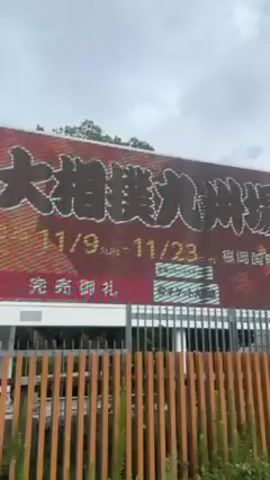 The video shows an outdoor scene with a large billboard advertising a sumo wrestling event in Kyushu, Japan. The billboard is predominantly red and features Japanese text and what appears to be an image related to sumo wrestling. A fence runs along the bottom of the billboard, with some greenery planted in front. People walk by in the foreground, including a man wearing a black baseball cap and a white face mask. In the background, a tall white building and a traditional Japanese building with a green roof can be seen amongst the trees. The sky is overcast.