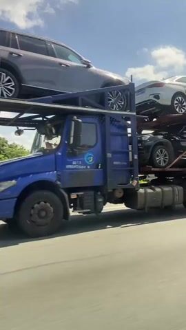 A car carrier truck is driving on a highway. On the top level of the car carrier, a dark-colored SUV is being transported. The truck is blue, and it has a logo on the side. The truck is driving on a paved road with multiple lanes. On the side of the road, there are trees and vegetation. The sky is blue with some white clouds. The camera appears to be mounted on another vehicle traveling alongside the truck.