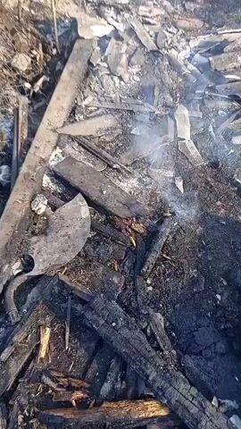 The video shows the aftermath of a fire. The ground is covered in debris, including charred wood, broken concrete, and crumpled metal. A structure in the background appears to be partially destroyed, with smoke still rising from it. The scene is littered with various building materials that are black and appear to have been burned. In the foreground, a person's legs, clad in dark pants and boots, are visible, suggesting someone is documenting the scene. The overall environment appears desolate and indicative of significant destruction.