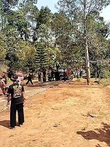 The video shows an outdoor gathering in a rural area with lush greenery and trees. In the foreground, a person wearing a black t-shirt with designs and black pants is walking away from the camera. Behind them, on the left, several people in traditional attire with elaborate headdresses are gathered, some appearing to be performing or preparing to perform. To the right of the performers, a dark-colored vehicle, possibly an SUV, is parked on a dirt road, with a group of people standing around it. Further in the background, a white pickup truck is visible under a carport attached to a house. The scene suggests a cultural event or celebration taking place in a residential area. The overall atmosphere is lively, with several individuals engaged in various activities.