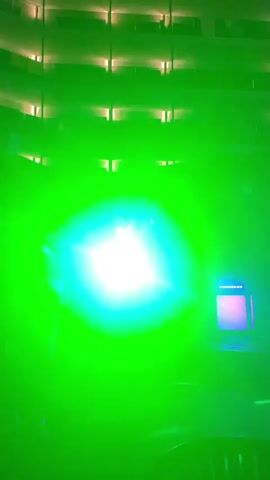 The video displays a stage with colored lights. In the first frame, a green light illuminates the stage, casting a bright glow. In the second frame, the light changes to blue, creating a different ambiance. In both frames, the stage is set against a backdrop of what appears to be a multi-story building with illuminated windows. There are people visible on the stage, but they are indistinct due to the lighting. The overall impression is that of a performance or event taking place.