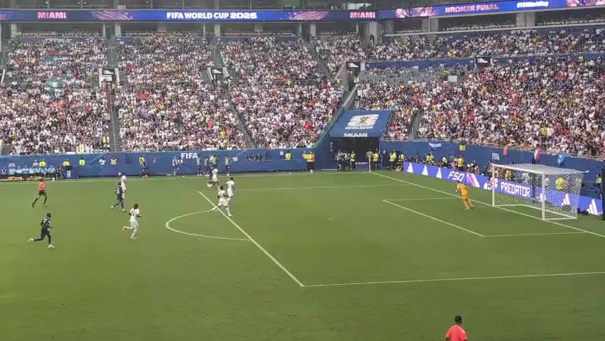 The video shows a soccer match from a high angle in a crowded stadium. The field is green, and the stands are filled with spectators in white and black attire. The game is in progress, with players in blue and white uniforms on the field. The ball appears to be near the center of the field, with players from both teams converging on it. A goal with netting is visible in the background, and a goalie in orange is positioned near it. A referee in a red shirt and black shorts is also visible on the field. The atmosphere seems lively, with some spectators in the foreground raising their hands.