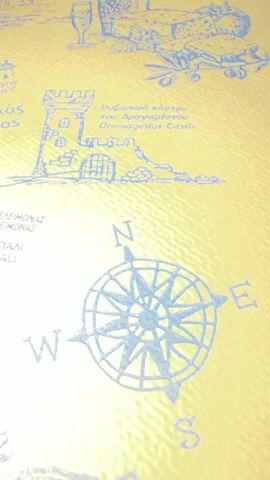 The video pans across a map printed on a textured yellow surface. The map depicts geographical features and landmarks in blue ink. Key elements visible include a compass rose with cardinal directions (N, S, E, W), a drawing of a castle labeled "Dramagestos Castle", various place names in Greek and English such as "Astakos" and "Ag. Panteleimonas", and small illustrations of buildings, animals, and a bottle and glass. The camera movement is smooth, suggesting a deliberate exploration of the map's details.
