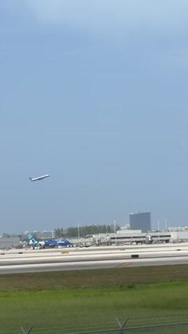 A commercial airplane is seen ascending into the sky against a bright blue sky with a few wispy clouds. In the background, an airport terminal with several parked airplanes, including one with blue and white livery, is visible. A tall building is also situated in the distance. The foreground shows a grassy area and a chain-link fence. The airplane is in the upper left portion of the frame and is traveling from left to right as it gains altitude.