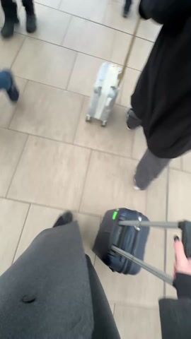 A person, dressed in a gray coat and dark shoes, is pulling a blue carry-on suitcase on a tiled floor. The camera is positioned from the person’s point of view, looking down at their feet and the suitcase. The person is walking past other people who are also pulling suitcases. The surroundings appear to be a train station or airport terminal. A sign is visible on the side that includes images of people. The camera angle frequently changes, including tilting up to show other pedestrians, before returning to the subject’s feet and suitcase.