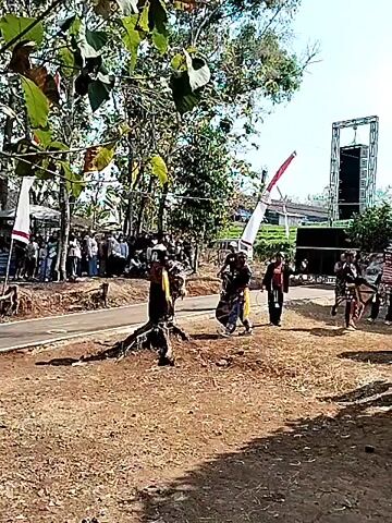 A group of performers are engaged in a ritualistic dance outdoors. The performers are wearing traditional costumes and masks, and some are carrying props such as swords and whips. They are performing in front of an audience, some of whom are standing and watching, while others are seated. There are also several trees and a stage set up in the background, suggesting this is some kind of outdoor festival or performance. The weather appears to be sunny and warm, and the overall atmosphere seems to be festive.