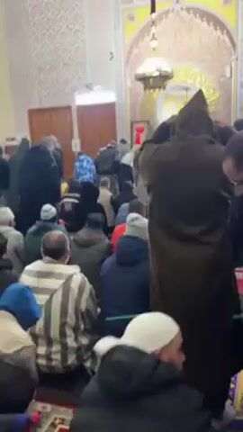 The video shows a large group of men inside what appears to be a mosque. They are standing or kneeling on a patterned rug. Some men are wearing hooded garments and casual clothing. The mosque features ornate tilework on the walls and ceiling, along with large pillars. A light fixture hangs in the center of the room. The men seem to be engaged in a religious activity, possibly prayer. The camera pans across the scene capturing the details of the interior and the individuals present.