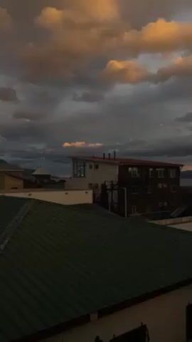 The video shows a view of a town at dusk. The sky is filled with dark gray clouds, highlighted by streaks of orange and gold from the setting sun. In the foreground are the rooftops of several buildings, primarily green and white. In the middle ground, a two-story building stands out with a brown facade and white trim, its windows reflecting the dim light. A black staircase is visible on its side. Beyond the buildings, a body of water stretches towards the horizon, where distant mountains are silhouetted against the remaining light of the sunset. The camera pans slowly to the right, revealing more of the cityscape and the body of water.