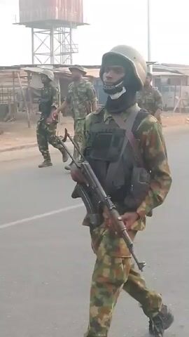 The video depicts several uniformed soldiers walking down a road. The first soldier is wearing a helmet and camouflage gear, and carrying a rifle. Two more soldiers, also in camouflage, follow behind. An ambulance with flashing red lights drives down the road in the opposite direction. There is a water tower and market stalls in the background. The weather appears hazy.