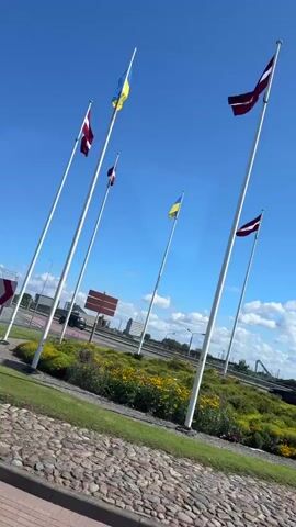 The video shows a series of flags flying on tall poles against a bright blue sky with scattered clouds. The flags appear to be the flags of Latvia (maroon and white) and Ukraine (blue and yellow). They are arranged in a semi-circle around a circular garden bed filled with green foliage and colorful yellow and purple flowers. In the background, there is a road with cars driving by and some trees. The ground in the foreground is paved with cobblestones. The overall impression is a sunny day with flags representing national pride or solidarity.