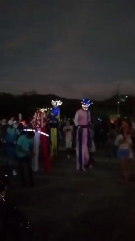 The video shows two people walking on stilts during a night event. They are surrounded by a crowd of people on the ground. The figures on stilts are wearing costumes and lit-up headdresses. The lighting is low, with streetlights and illuminated costumes providing visibility. The moon is visible in the dark sky. The camera is unsteady and moves with the crowd. The sound includes music or speech, which adds to the atmosphere of the event.