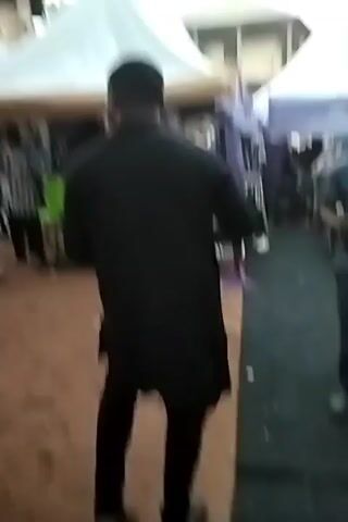 Here's a detailed description of the video:

The video shows an outdoor gathering, possibly a celebration or event, taking place under several white tents. The ground is partially covered with a black carpet, with sandy-colored dirt visible on the uncovered areas. There is litter scattered on the ground, including white paper.

The main subject of the video is a man seen from the back, wearing a long-sleeved black shirt and black pants. He is walking toward the tents. To his right, another man walks by, wearing a baseball cap, a patterned grey shirt, and dark pants. This man appears to be carrying several items in his hands.

Other people are visible in the background, some seated and some standing, under the tents.
