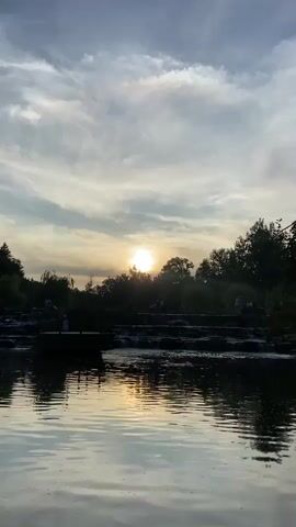 A video captures a sunset over a body of water, possibly a river or lake, with a series of small waterfalls cascading over rocks in the mid-ground. The sun, a bright orb, is visible in the sky, partially obscured by trees and clouds. The sky is a mix of blue and grey with wispy white clouds. The water is dark and reflects the sky and the sun's light. On the left side of the frame, there is a small wooden platform or dock extending into the water, with several silhouetted figures standing on it. Further back, along the edge of the water and near the waterfalls, more silhouetted figures are scattered, appearing to be enjoying the scenery. Lush green trees form a dense backdrop behind the waterfalls and along the water's edge. The overall impression is a peaceful, natural scene during golden hour.