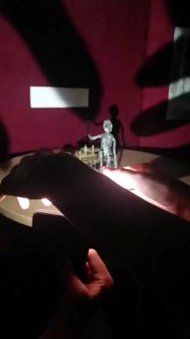 The video shows a silhouette of a skeleton figure and its shadow on a maroon wall. The skeleton is seated at a table with what appear to be chess pieces. There are also abstract shadows projected onto the wall behind the skeleton, one rectangular and white, and another that is more organic in shape. In the foreground, out of focus and silhouetted, are two pairs of hands. One pair of hands holds a smartphone, with the screen illuminated. The other hands are near a circular, illuminated surface, possibly a light table or stage. The scene suggests a shadow puppet performance or animation being created.