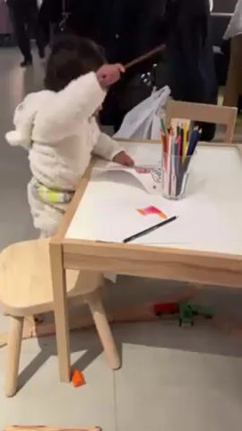 A toddler stands at a small, white table with a wooden frame, drawing with colored pencils on a piece of white paper. The child is wearing a light-colored hooded jumpsuit. A glass of colored pencils sits on the table along with loose pencils and the child's drawing. A small wooden chair is positioned next to the table. Underneath the table, a wooden train track is visible with a few toy trains. Other people are walking in the background, but they are out of focus. The floor is a light gray tile.