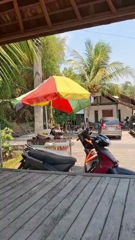 A man stands behind a food cart under a multicolored umbrella. The cart has signs that appear to be in Indonesian. Two motorcycles are parked in front of the cart. The background shows trees, a building, and a car. The lighting suggests it is daytime. The man appears to be preparing food.