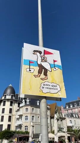 A sign depicting a cartoon goose in golf attire, with the words "BEACH GOLF DE TROUVILLE" below it, is mounted on a metal pole. The sign is weathered and appears to be made of wood. In the background, there are several buildings with distinctive architecture, including a domed building on the left and a pink and white striped building on the right. People are seated at tables outside a cafe or restaurant in front of the buildings. The sky is clear and blue, and a few birds are visible flying in the distance. The scene appears to be a sunny day.