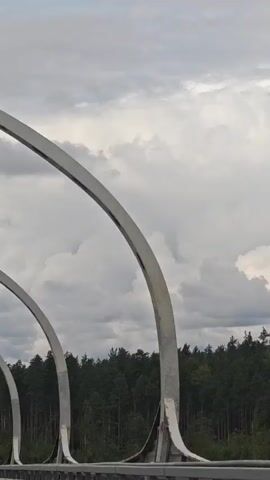 The video shows a perspective from a vehicle moving along a road. The road has a metal barrier on the right side and a series of white curved metal structures overhead, forming a tunnel-like effect. Beyond the barrier and structures, there is a dense forest of dark green trees against a cloudy sky. The camera angle is slightly tilted upwards, capturing the sky and the tops of the trees. There are no people visible in the video. The lighting suggests it is daytime, but the clouds indicate overcast weather.