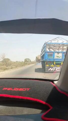The view from inside a moving Peugeot car shows the road ahead. The car is traveling on a paved road, and a large truck is moving ahead of it. The truck is blue and has a large tarp covering its cargo. The word "CONGRAT" is written on the back of the truck. The landscape surrounding the road is dry and grassy with scattered trees. The car’s dashboard is visible in the foreground.