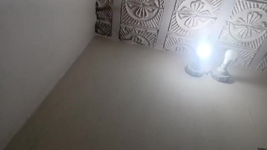 The video presents a static, upward-angled view of an indoor room, focusing on a corner where a decorative ceiling meets a plain wall. The ceiling features a grid of light-colored, square tiles, each embossed with a circular, floral-like pattern and radiating lines. Some of these tiles, particularly near the corner, show imperfections or an unfinished appearance. Two bright, white lights, likely LED bulbs, are mounted on the ceiling, strongly illuminating the area. The walls are painted a uniform white or off-white. No people or activities are visible. The setting is an indoor space located in Kakki, Pakistan.