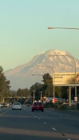 The video shows a view from inside a car driving on a road. In the distance, a snow-capped mountain looms large against a clear sky. The sun appears to be low in the sky, casting a warm glow. There are other cars on the road, and streetlights are visible. A billboard on the right side of the road advertises "Grow your everyday getaway" with the Watson's logo. The overall scene suggests a scenic drive towards a majestic natural landmark.