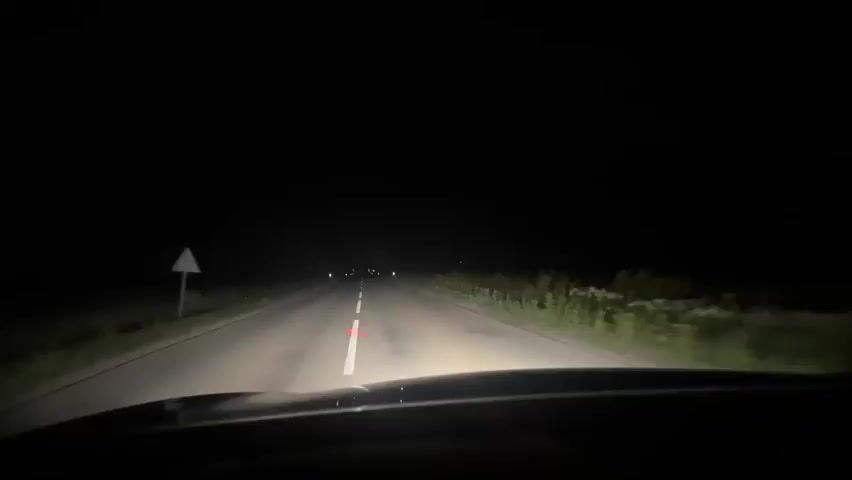 The video shows a first-person perspective of driving a vehicle at night on a paved road. The headlights illuminate the road ahead, which has a white solid line on the left and a dashed white line to the right. The surrounding environment is dark, with faint lights visible in the distance and some vegetation on the sides of the road. The sky is black. There is a triangular road sign on the left side of the road.