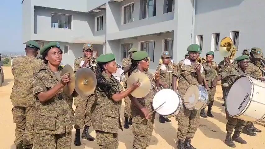 A military band composed of women in camouflage uniforms and green berets is playing in front of a modern building. The band members are holding and playing various instruments, including cymbals, snare drums, and brass instruments such as tubas and trumpets. Some women are actively striking cymbals, while others are playing drums or holding their instruments ready. The building appears to be a light grey, multi-story structure with multiple windows. The ground is a dirt or gravel surface. The overall atmosphere suggests an outdoor ceremony or event.