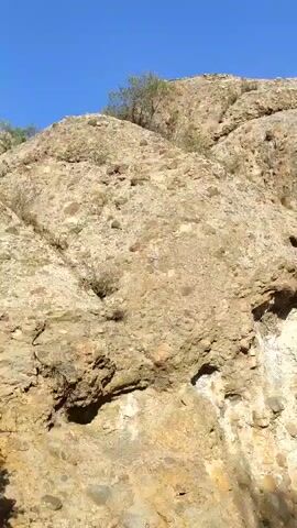 The video shows a rocky, arid landscape under a clear blue sky. The camera pans across a large, weathered rock formation that appears to be part of a hillside. The rock is composed of various shades of brown and tan, with visible pebbles and stones embedded within its structure. Some sparse vegetation, consisting of dry grasses and small bushes, grows on the slopes. The terrain is uneven and appears to be a natural, undeveloped area.