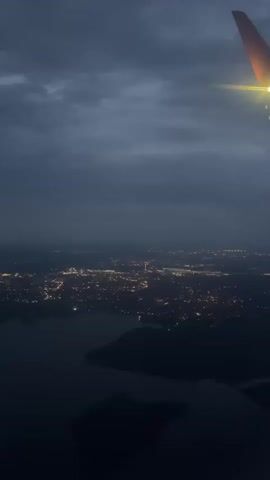 The video shows an aerial view of a city at dusk. The city lights are visible as small yellow and white dots scattered across the landscape. A body of water, possibly a lake or river, is visible in the foreground, with dark landmasses silhouetted against it. The sky is dark and cloudy, with a hint of orange light on the horizon, suggesting either sunrise or sunset. A portion of an airplane wing, with its navigation light on, is visible in the upper right corner of the frame. The overall atmosphere is serene and tranquil, with a sense of approaching night.