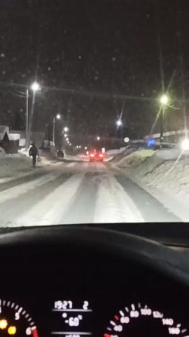A car is driving at night on a snowy road. Streetlights illuminate the road, and there are snowbanks on either side. A person is walking on the left side of the road, and a car is ahead. The dashboard of the car shows the time is 19:27 and the temperature is -60 degrees. Music is playing.