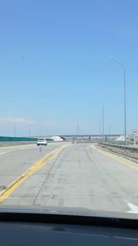 The video shows a first-person perspective from inside a car driving on a highway. The road is paved with concrete and has yellow and white lane markings. There are streetlights visible along the sides of the road. In the distance, there is an overpass or bridge. Several cars are visible on the road, with one white car in front of the camera. There are also various road signs, including directional signs with arrows. The sky is clear and blue, suggesting a sunny day. The overall environment appears to be a modern highway in an open area.