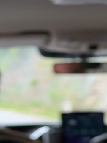 The video shows the interior of a car, looking forward through the windshield. The scene outside is blurred due to motion, but appears to be a mountainous or hilly area with vegetation. There is a rearview mirror reflecting a person's head and shoulders. A dashboard display screen is visible in the lower part of the frame, showing a purple interface with circular icons. The time stamp at the beginning of the video is 00:00, and the time on the dashboard screen reads 10:01. The overall impression is that of a car journey in a scenic, possibly rural, location.