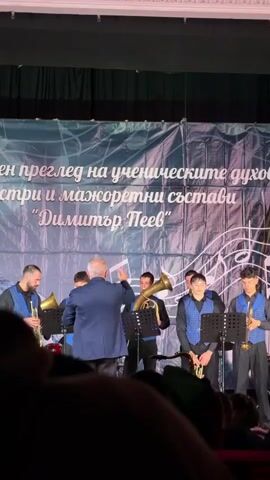 The video shows a brass band performing on a stage in front of an audience in a theater. The band consists of about eight members, including the conductor, who is wearing a blue suit and directing the music. The banner behind the band indicates that the event is a review of student wind orchestras and majorette bands. The theater has red seats and interior decorations, and the lighting on the stage is bright. The audience is visible in the foreground, consisting mostly of silhouettes of people sitting and watching the performance.
