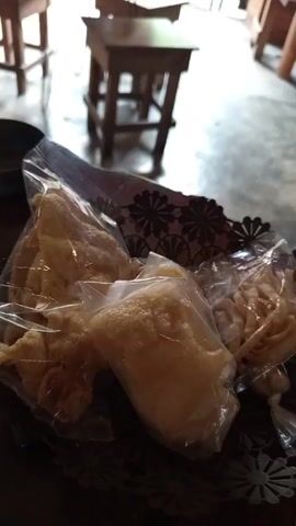 The video shows a variety of Indonesian crackers, likely Kerupuk, individually packaged in clear plastic bags and arranged in a dark, decorative metal basket. The crackers vary in shape and texture; some are large and flat, others are smaller and bubbled, and some appear as twisted shapes. The basket is placed on a dark surface, possibly a table. In the background, two wooden stools are visible on a light gray floor. The scene is indoors, and the lighting seems natural.