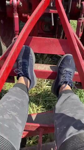 The video shows a close-up of a red agricultural implement being used to till the soil. The implement appears to be a disc harrow, with several rotating discs visible. The camera is positioned from the perspective of someone standing on the implement, holding onto a red metal bar. The person's hand, wearing a black watch, is visible in the foreground, along with their leg wearing camouflage pants and a dark blue sneaker. The tractor's large tire is also partially visible in the upper left corner. The ground is a mix of soil and grass. The footage shows the implement digging into the earth, churning up soil and vegetation. The video is short, with the camera angle shifting slightly throughout, offering different views of the tilling process and the machinery. The lighting suggests it is daytime, with bright sunlight.