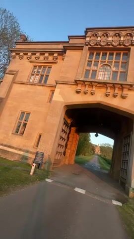 The video shows a forward-facing view of a long driveway as seen from under a large, stone archway with open wooden gates. The driveway is asphalt and leads away from the viewer toward a distant building. The archway appears to be part of a larger stone building with multiple windows visible on the structure above. A street light is visible in the middle of the archway. The sun appears to be setting or rising, casting warm light on the stone structure and the surrounding greenery along the driveway. Two signs are visible on either side of the archway entrance, though their contents are not legible.